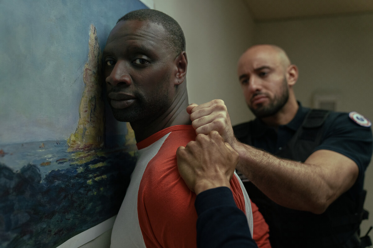 Um homem negro vestindo camisa branca e laranja é pressionado contra a parede por um policial branco, de barba curta e cabelos raspados, que o segura pela roupa