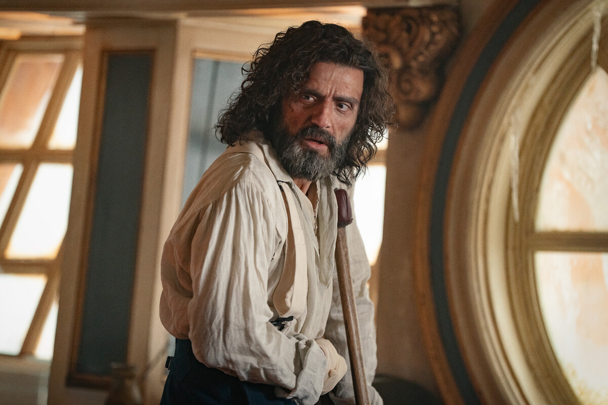 A bearded man with long hair, wearing a loose shirt, stands indoors by a large round window on a ship. The room is warmly lit with ornate wood decor. The mood is tense and dramatic.
