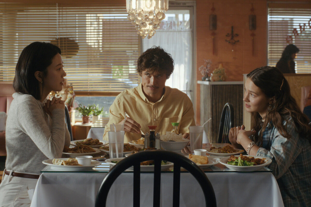 Three people are sitting at a table in a cozy restaurant, having a meal together with plates of food, drinks, and chopsticks, engaged in conversation under a chandelier with sunlight streaming through window blinds.