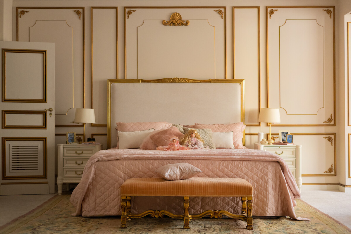 Um quarto amplo com paredes brancas adornadas com dourado tem uma cama em seu centro, com um pequeno sofá na ponta e uma cômoda de cada lado da cama