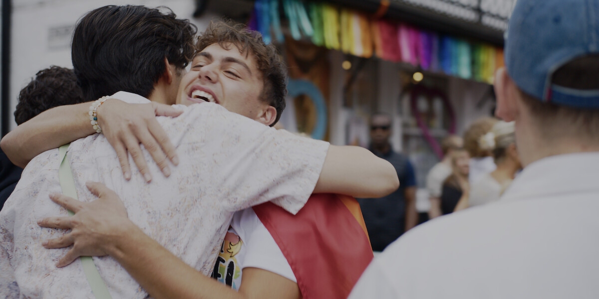 Two people hugging and smiling at an outdoor event with a rainbow banner in the background, suggesting a celebration or pride festival, surrounded by other attendees.