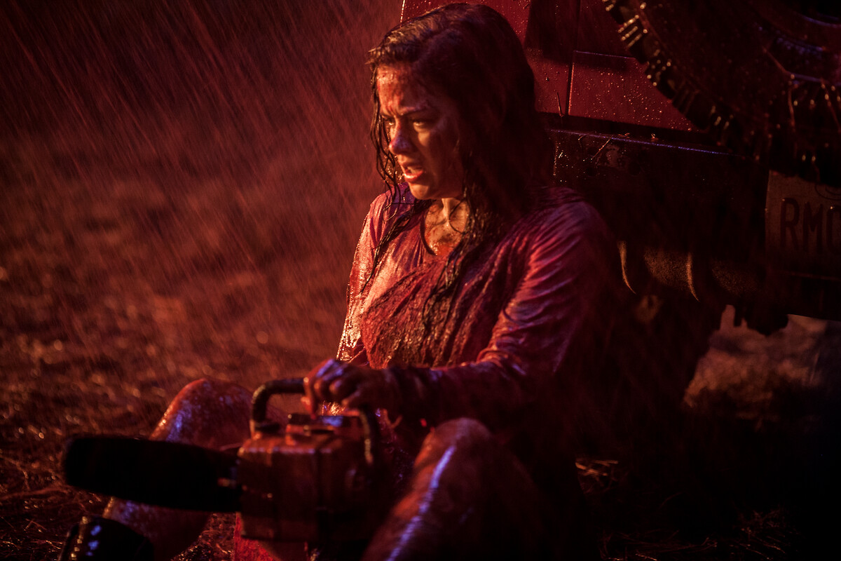 A woman covered in blood sits on the ground in the rain under a red light, leaning against a vehicle while holding a chainsaw, appearing exhausted in a tense, dramatic outdoor night scene.