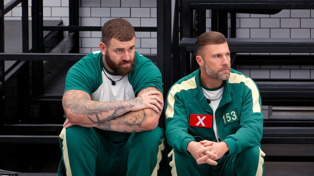 Two men in green tracksuits with numbers sit on a bench in a stark, tiled room, appearing pensive and tense, suggesting a competitive or high-stress environment.