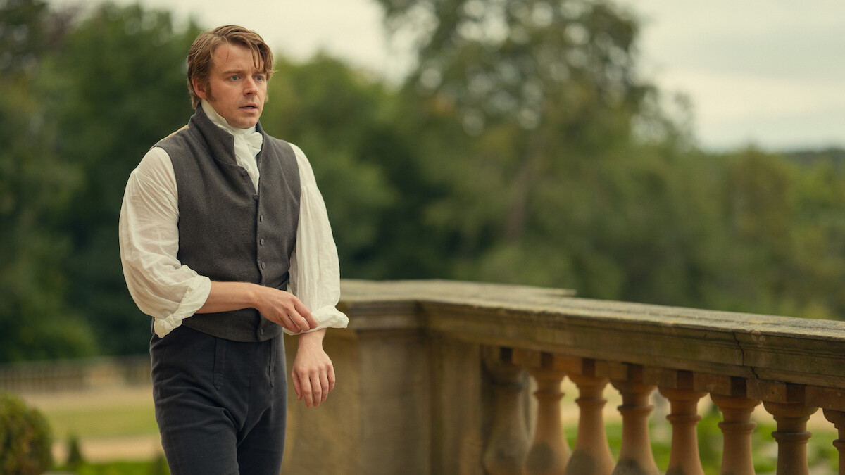 A man in period clothing stands outside on a stone terrace with ornate railing, surrounded by greenery and trees, giving a historical or vintage atmosphere.