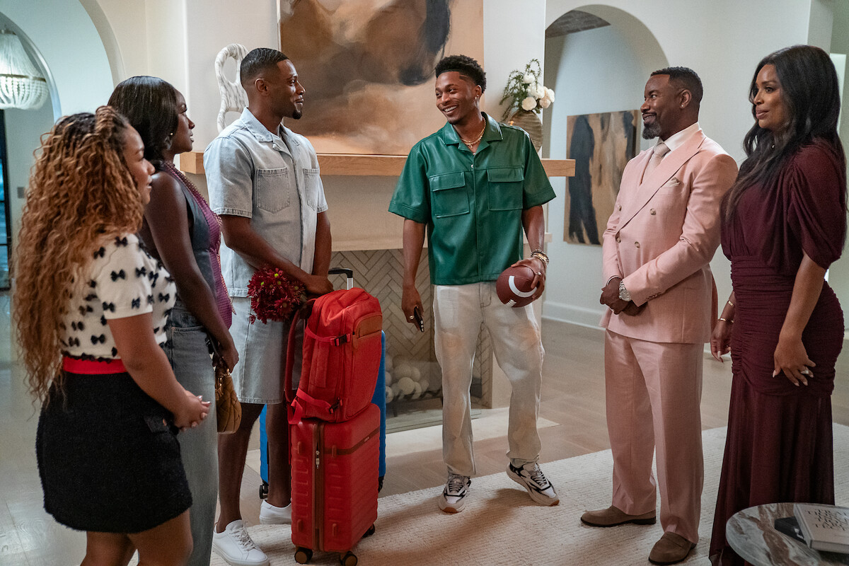 Six people gather in a stylish, modern living room. One person holds a football and luggage, suggesting arrival or departure, while others smile, dressed in a mix of casual and formal outfits. Bright, elegant home interior.