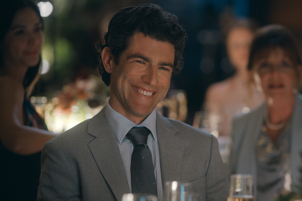 A man in a suit smiles while sitting at a formal event or celebration, surrounded by other well-dressed guests and soft lighting, with drinks and flowers visible on the table.