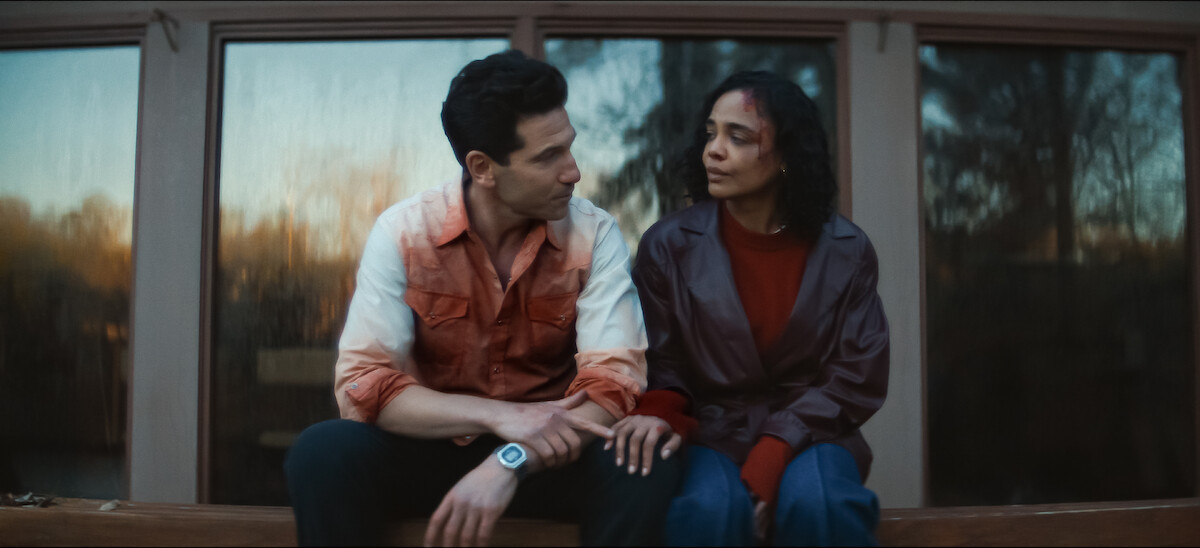 A man with blood on his shirt and woman sit closely on a wooden bench outside a building at sunset. The background shows large glass windows reflecting trees.