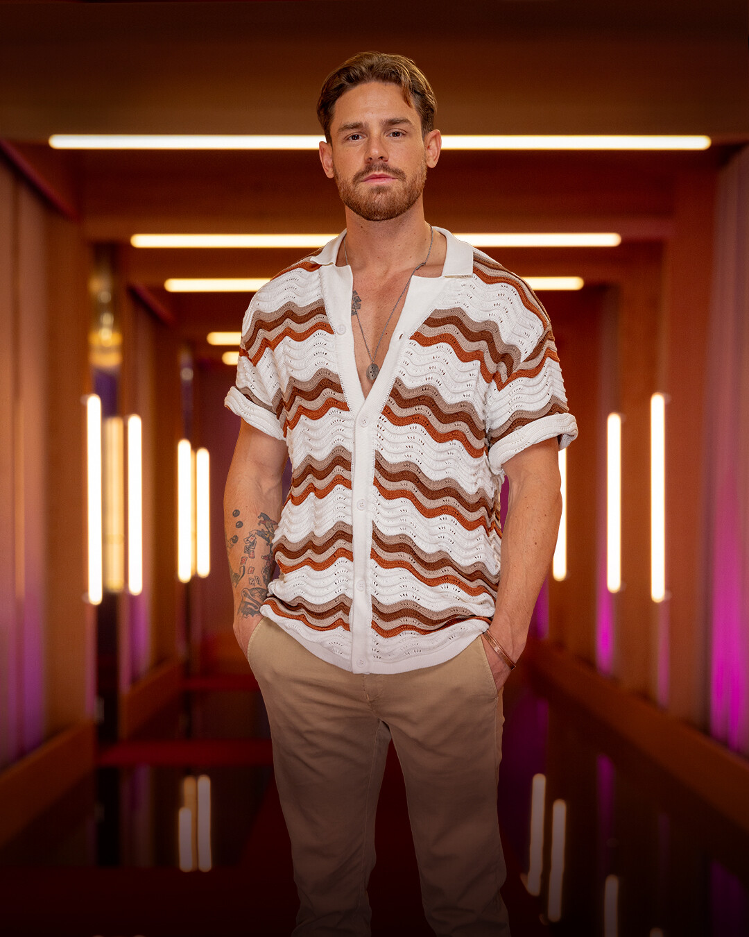 Man with light brown hair and a beard, wearing a white and brown wavy-patterned shirt and beige pants, standing in a modern hallway with warm lighting and neon accents.