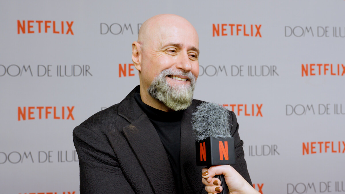 Um homem branco, careca e com barba grisalha dá entrevista em frente a um microfone com a canopla da Netflix