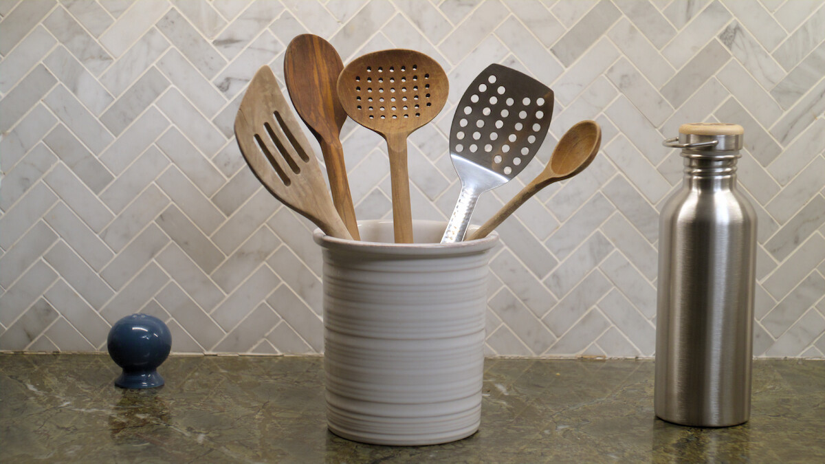 Kitchen countertop with a ceramic holder containing assorted wooden and metal cooking utensils, a stainless steel water bottle, and a blue salt shaker in front of a herringbone tile backsplash.