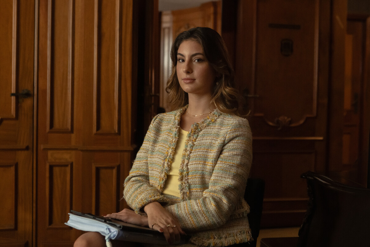 Mulher branca veste um casaco listrado e segura uma encadernação em frente a uma parede revestida de madeira