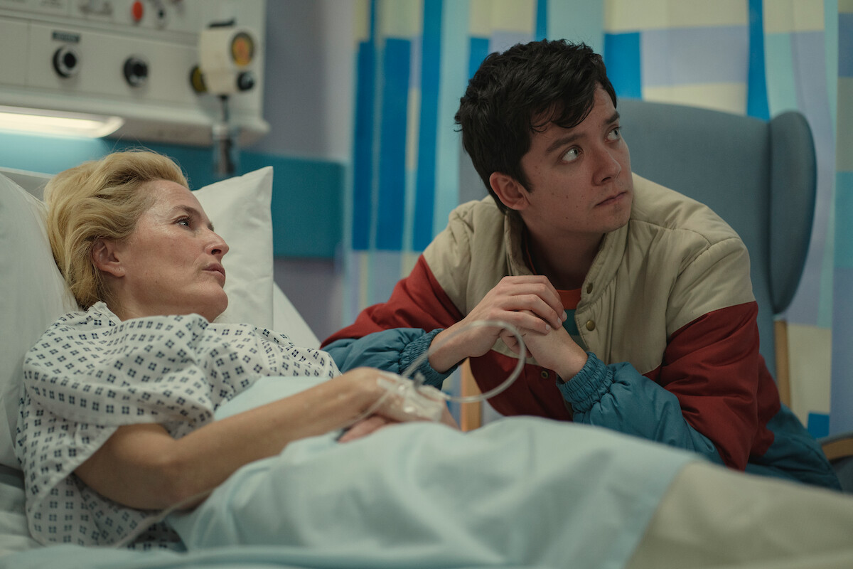 Gillian Anderson as Dr. Jean Milburn lays in a hospital bed holding hands with Asa Butterfield as Otis Milburn in Season 3 of ‘Sex Education.’