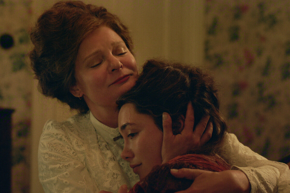 Two women in vintage clothing share an emotional embrace in a warmly lit, floral-wallpapered room, suggesting comfort and support in a historical or period setting.