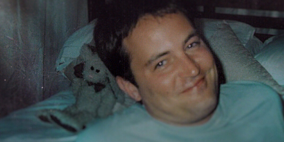Smiling man lying on a bed with white pillows and a teddy bear in the background, in a softly lit bedroom environment.