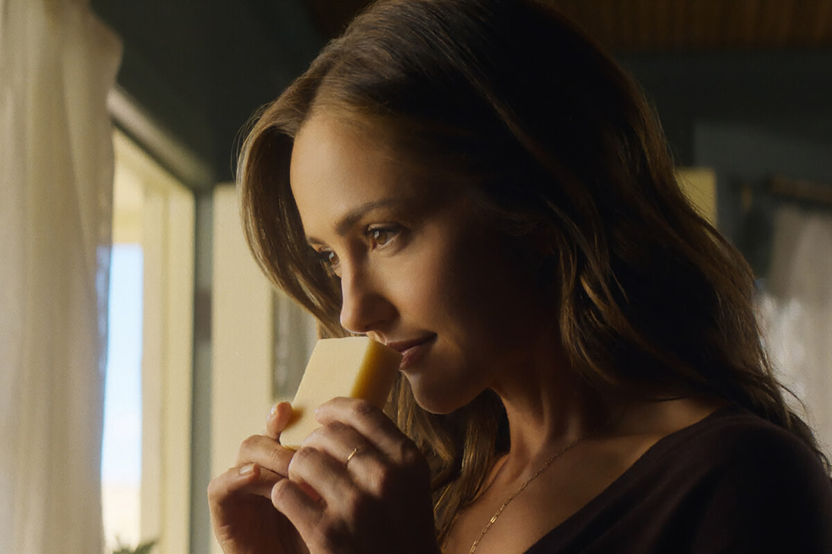 A woman stands indoors by a sunlit window, holding and smelling a piece of cheese, with soft natural light highlighting her face and a calm, cozy atmosphere surrounding her.