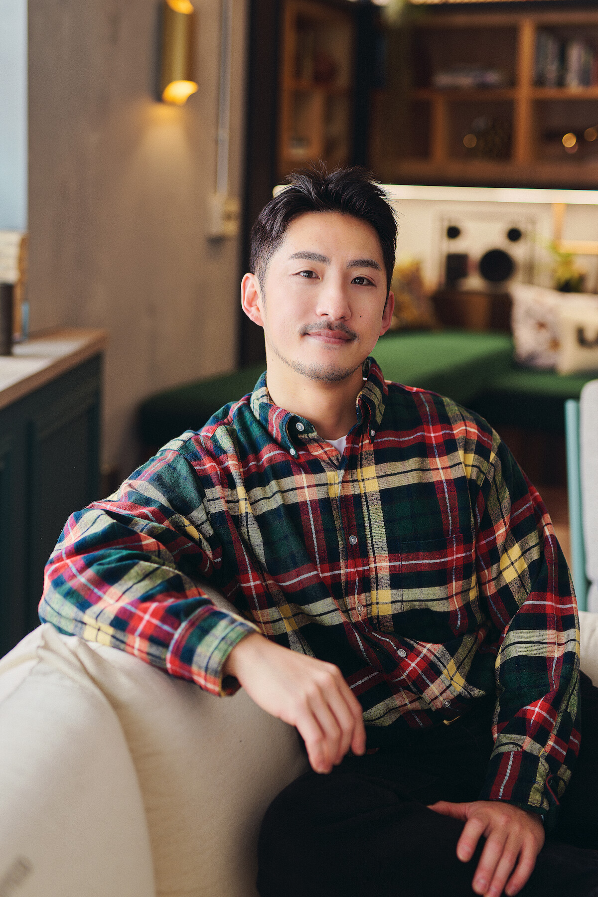 Man in a plaid shirt sitting relaxed on a sofa in a cozy, warmly lit living room with shelves, decor, and soft lighting in the background, creating a comfortable and inviting atmosphere.