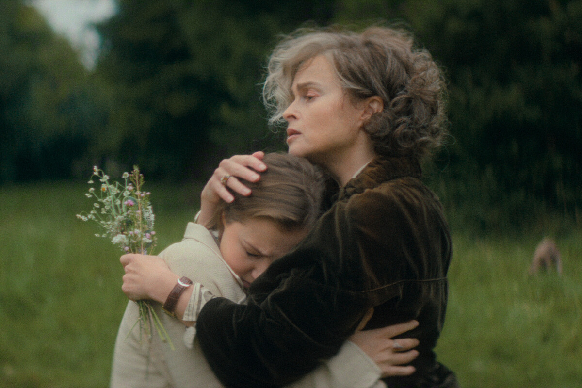 Woman with gray hair in dark coat hugs a sad young girl holding wildflowers in a grassy outdoor field. Both appear emotional, suggesting comfort and support in a natural, green environment.