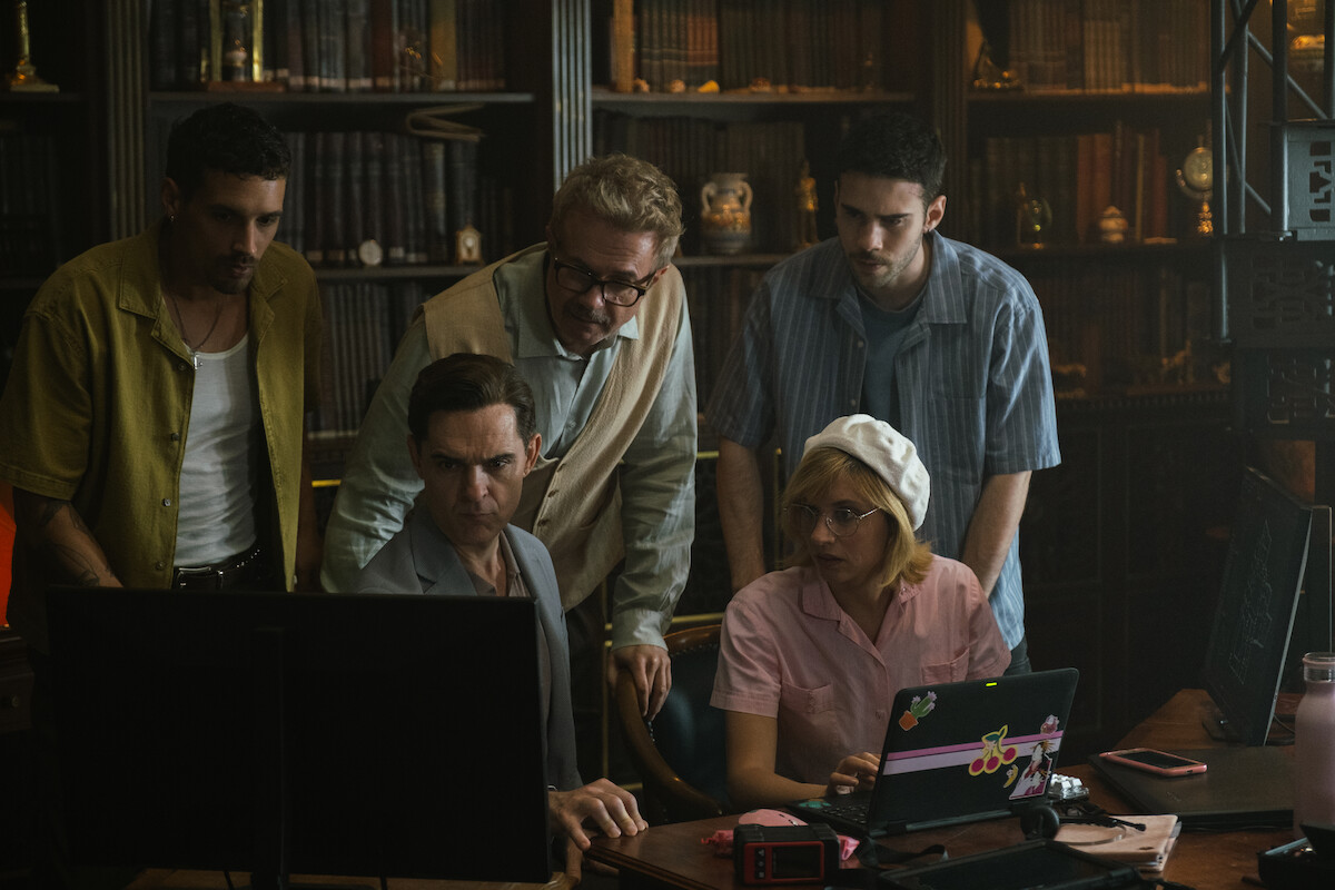 Five people in a library or study room, gathered around two computer monitors with bookshelves in the background, appearing focused and engaged in collaborative work or problem-solving.