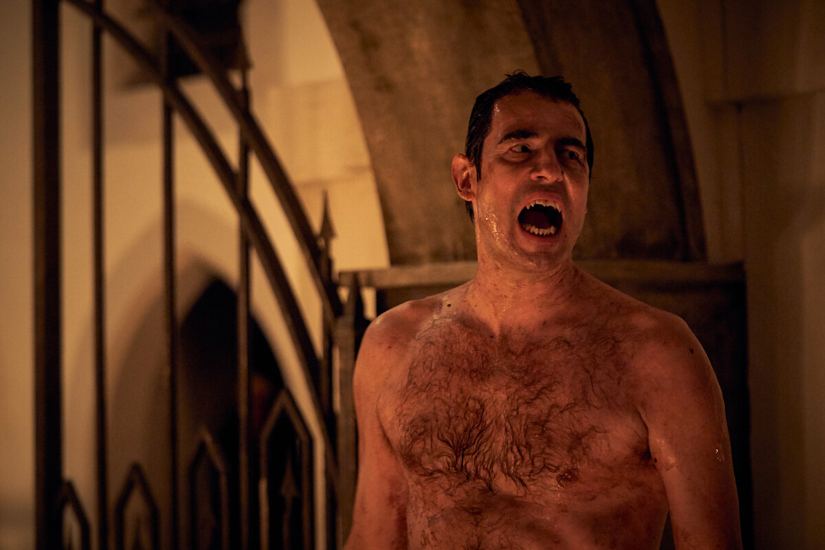 Shirtless man with wet hair stands in a dimly lit Gothic setting, mouth open as if shouting, with wrought iron gate and stone arch in the background, creating a dramatic and tense atmosphere.