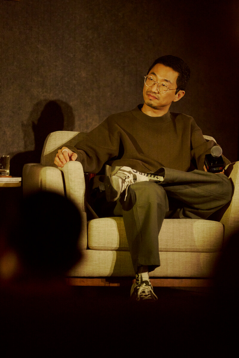 A man wearing glasses and casual clothing sits on a beige armchair on stage, holding a microphone. The background is dark and soft lighting highlights him, suggesting a panel discussion or interview setting.