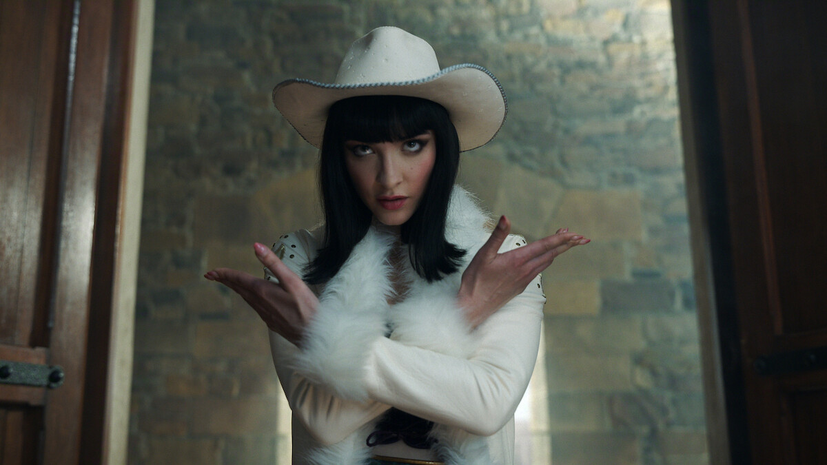 Lera Abova as Miss All Sunday, wearing a white cowboy hat and white fur-trimmed outfit stands indoors, in front of a stone wall, crossing her arms in an X gesture, between two wooden doors, with dramatic lighting.
