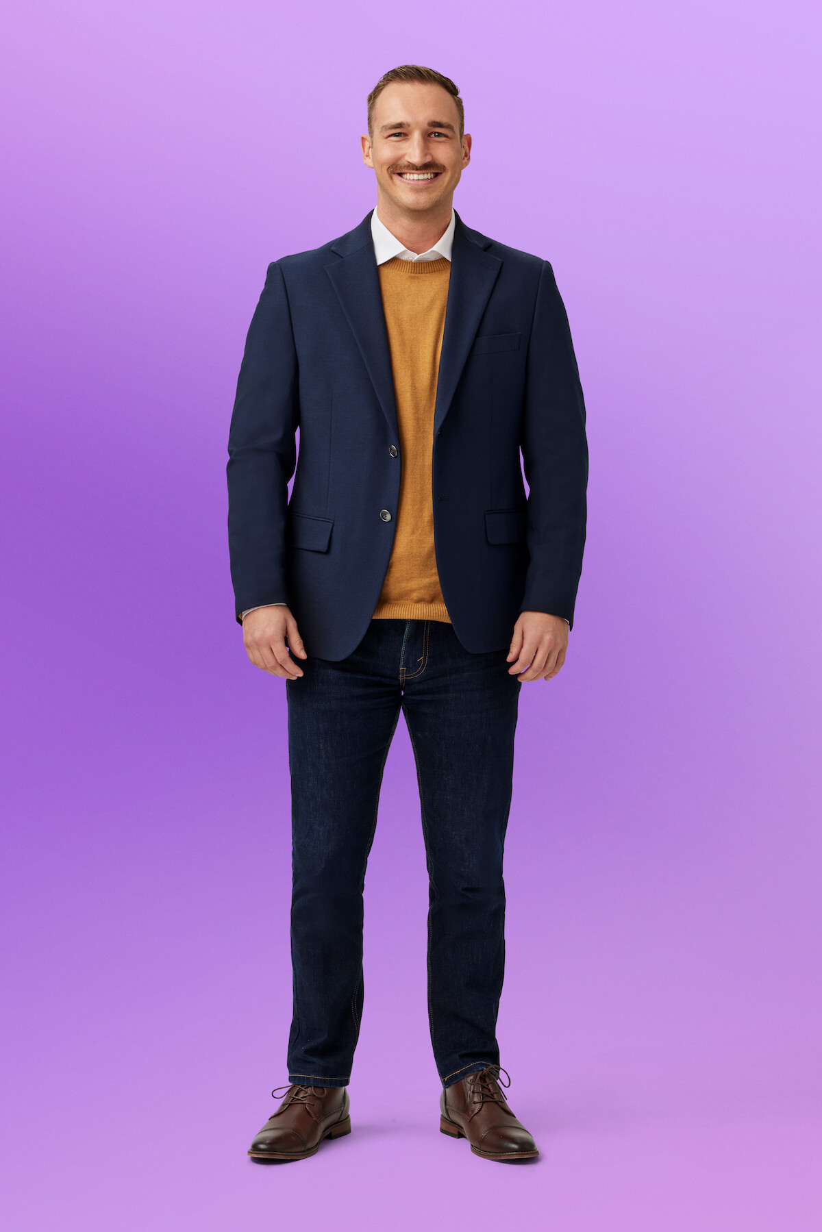 Brennan wearing a blue blazer, mustard sweater, white shirt, dark jeans, and brown shoes, standing against a purple gradient studio background.