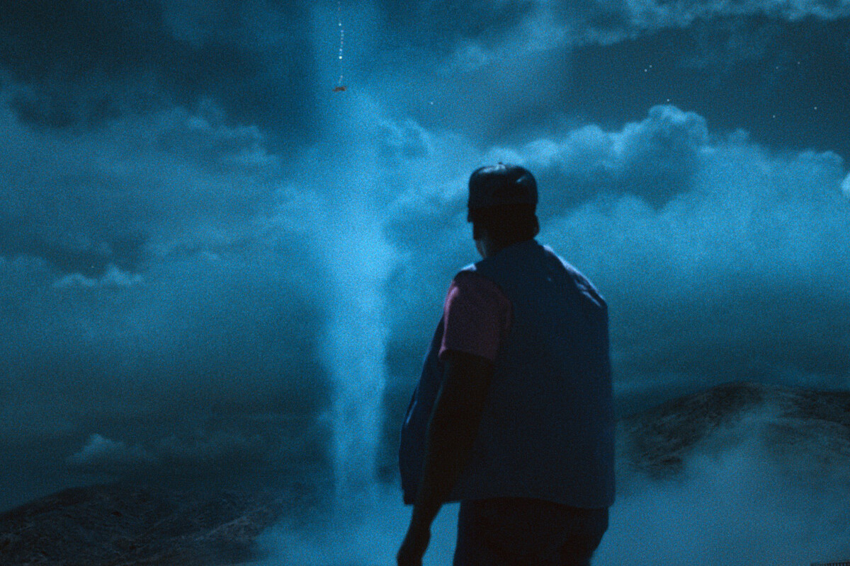A person stands outdoors at night under a cloudy sky, looking up at a bright beam of light extending into the sky with an object levitating high above, surrounded by rolling hills and mist.