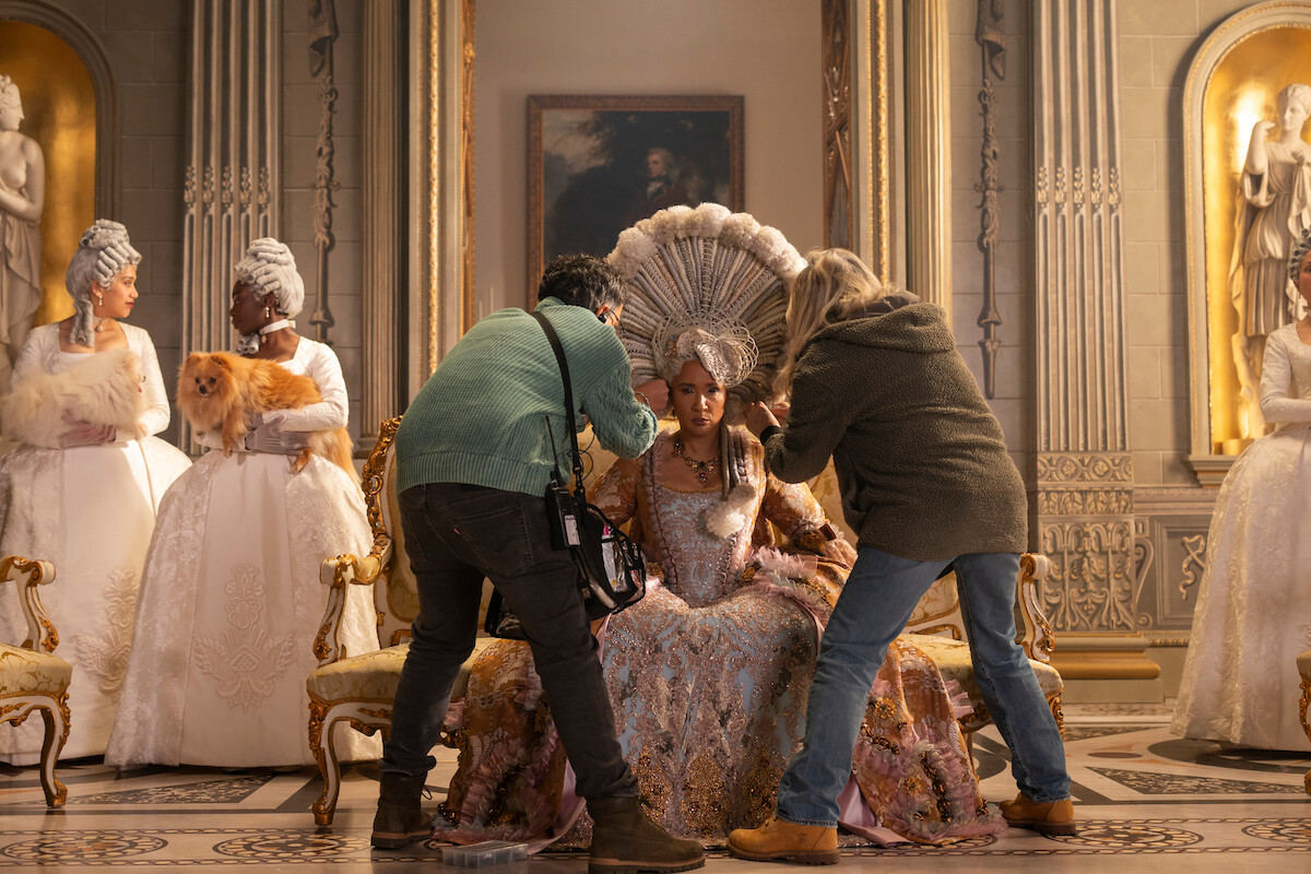 Integrantes do elenco com figurinos de época elaborados se preparam em um cenário suntuoso de palácio. A equipe ajusta o cabelo e a roupa da atriz principal, enquanto, ao fundo, outras pessoas de peruca e vestidos ornamentados aguardam, uma delas segurando um cachorro peludo.
