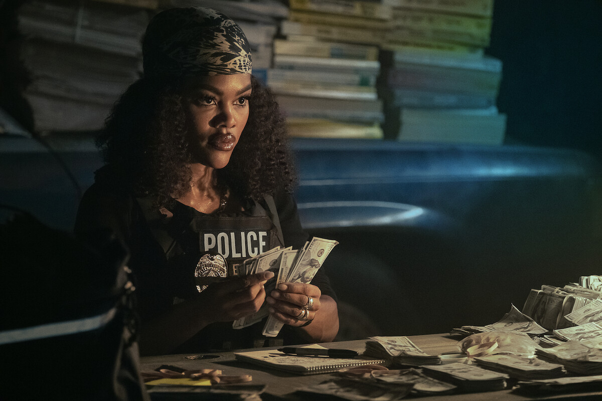 A police officer sits at a desk in a dimly lit room counting cash, with stacks of documents and evidence bags spread out.