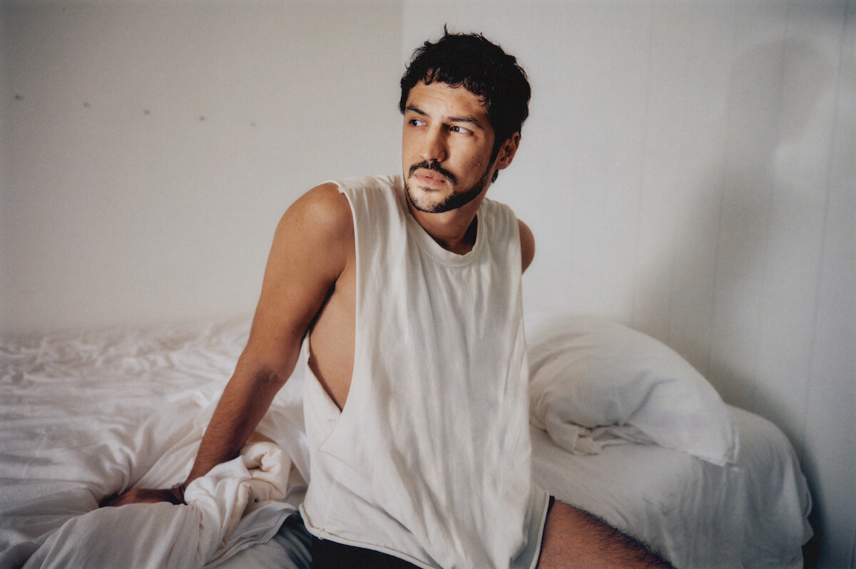 Gabriel Leone wears a white tank top and sits on a white bed.