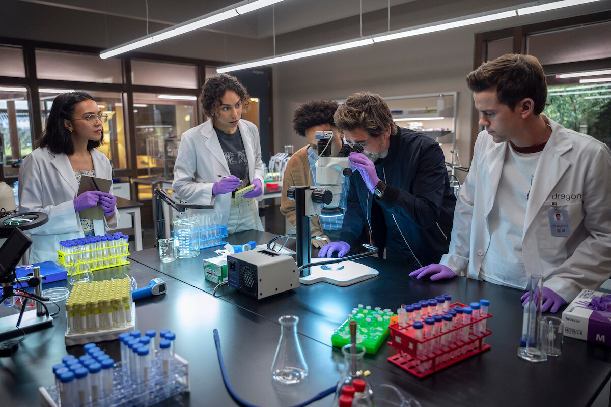The Dragon team stands around a lab table as Ellis Dragon (Rob Lowe) peers into a microscope.