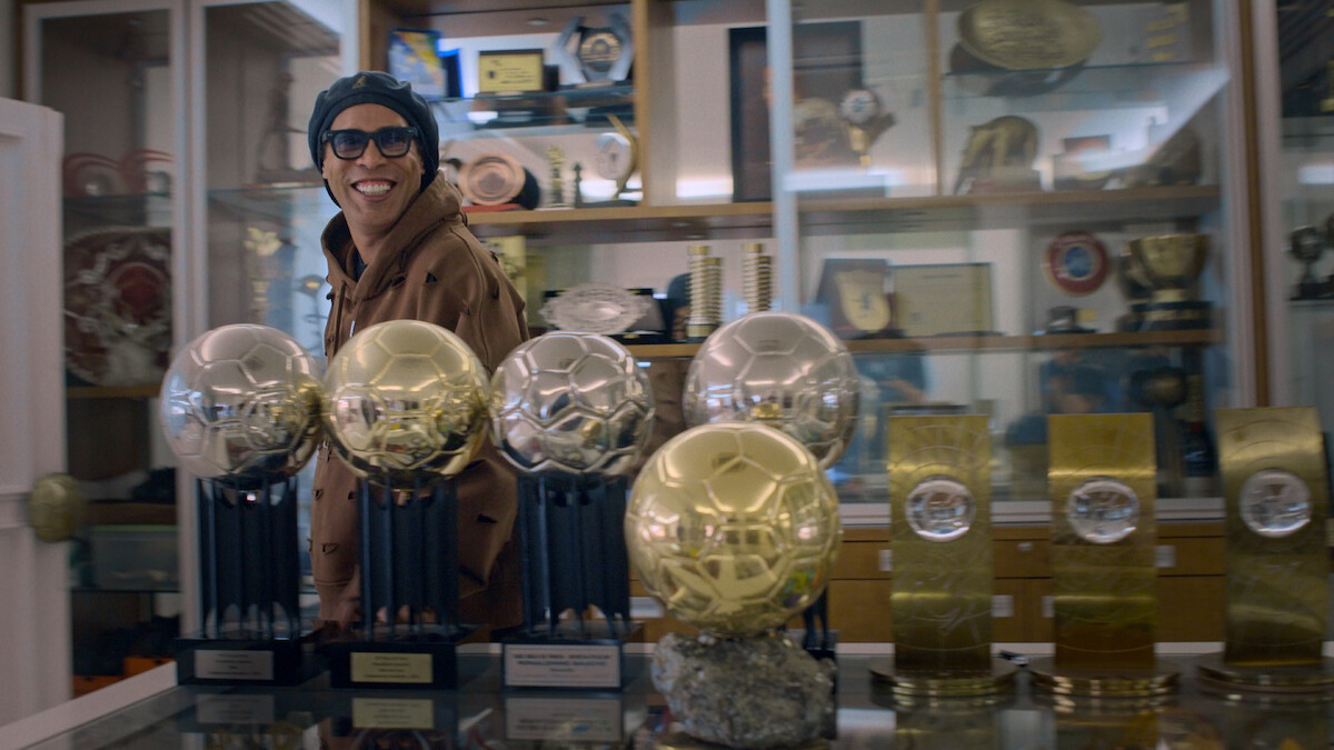 Ronaldinho Gaúcho smiling and wearing glasses stands in a trophy room filled with numerous awards and medals displayed in glass cases and on shelves, with several golden soccer trophies prominently on a table in the foreground.