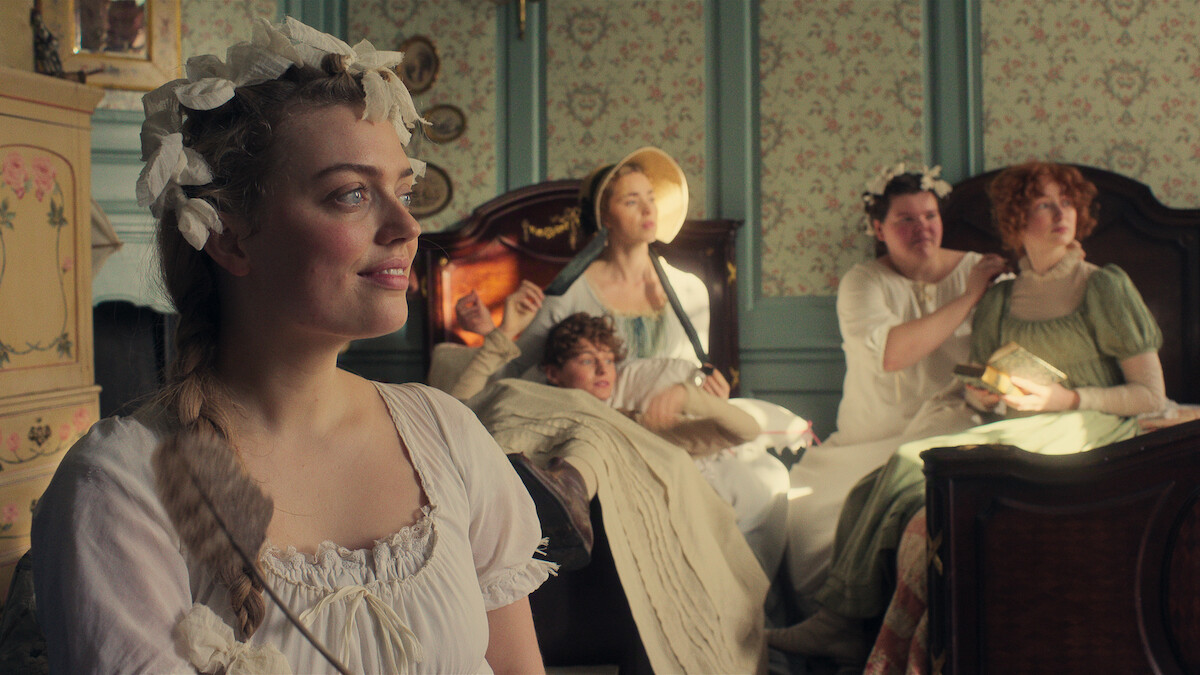 Five women in period clothing sit and relax in a vintage, pastel-colored bedroom with floral wallpaper and wooden beds, bathed in natural light, creating a cozy and historical atmosphere.