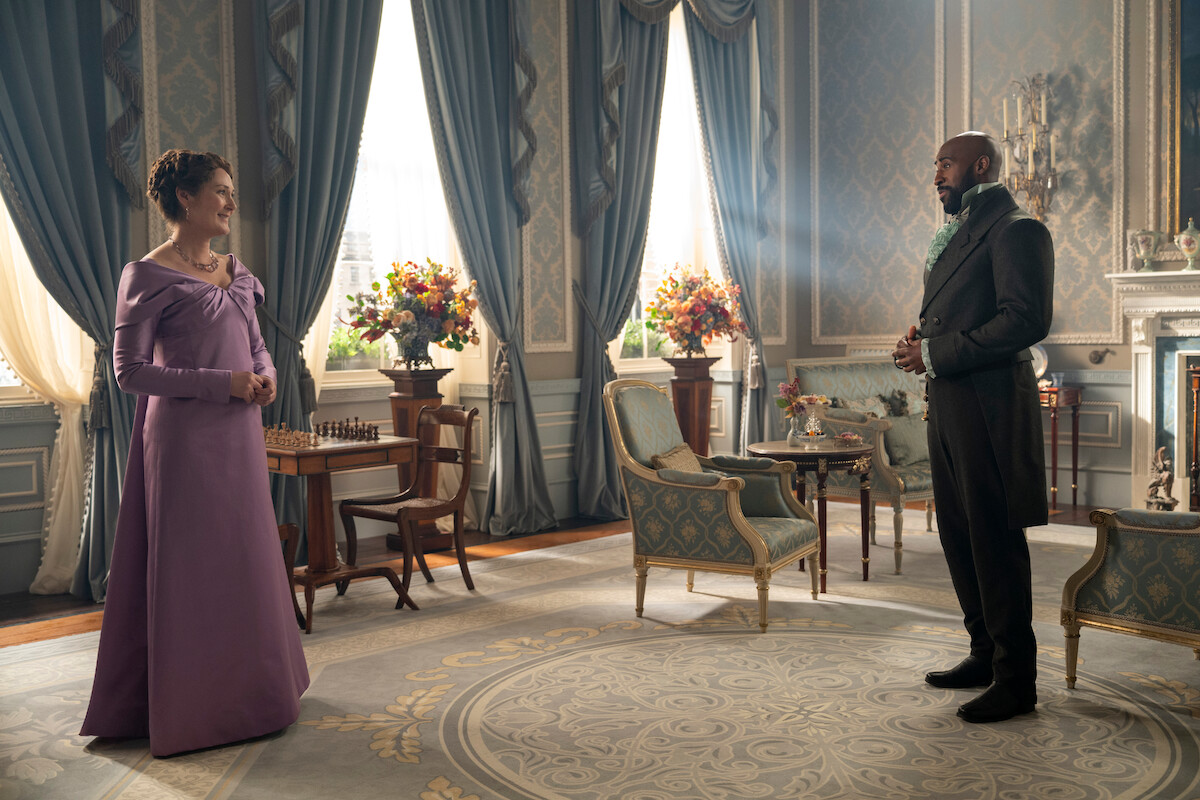Uma mulher de vestido roxo e um homem com roupa formal em pé, olhando um para o outro, em um salão com iluminação natural e móveis, janelas grandes, flores e decoração clássica.