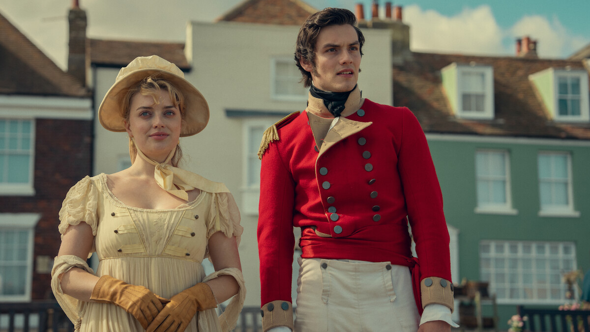 Two people in Regency-era costumes stand outdoors in front of historic houses; the woman wears a pale dress with gloves and bonnet, the man wears a red military uniform, both looking ahead under a partly cloudy sky.