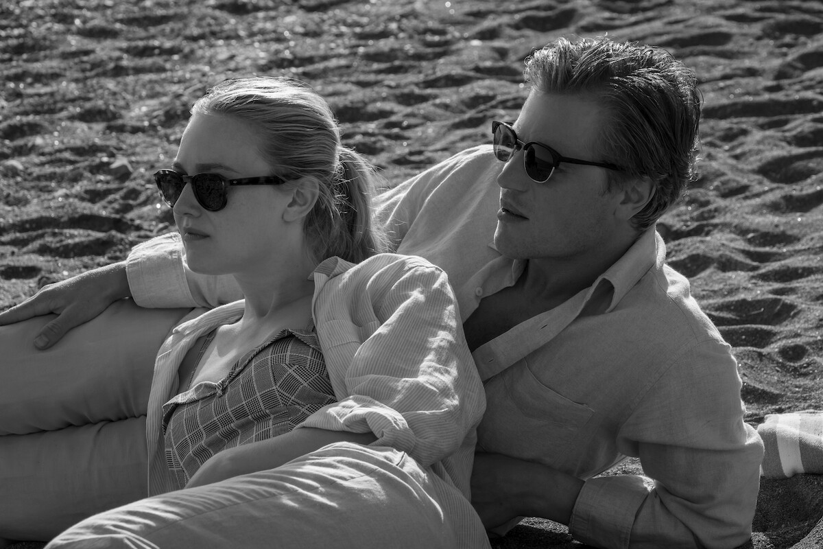 Black-and-white photo of Dakota Fanning as Marge Sherwood and Johnny Flynn as Dickie Greenleaf sitting on a beach together in ‘Ripley’