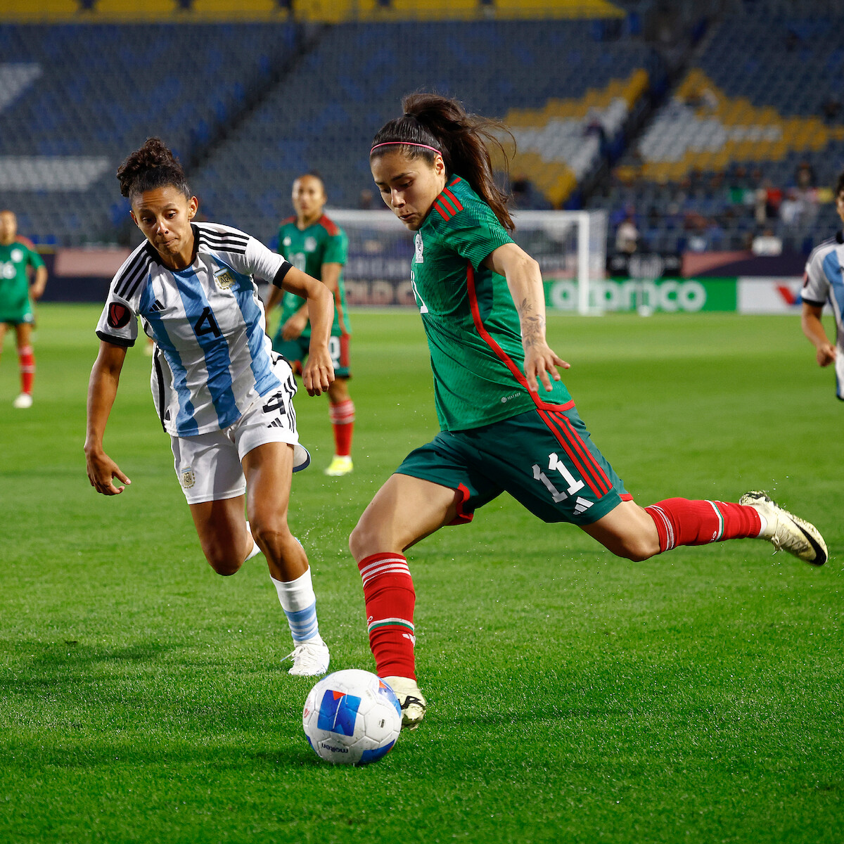 Two female soccer players compete for the ball during a match in a stadium, with one player in a green and red uniform about to kick the ball and another in a white and blue uniform defending, while teammates and spectators watch in the background.