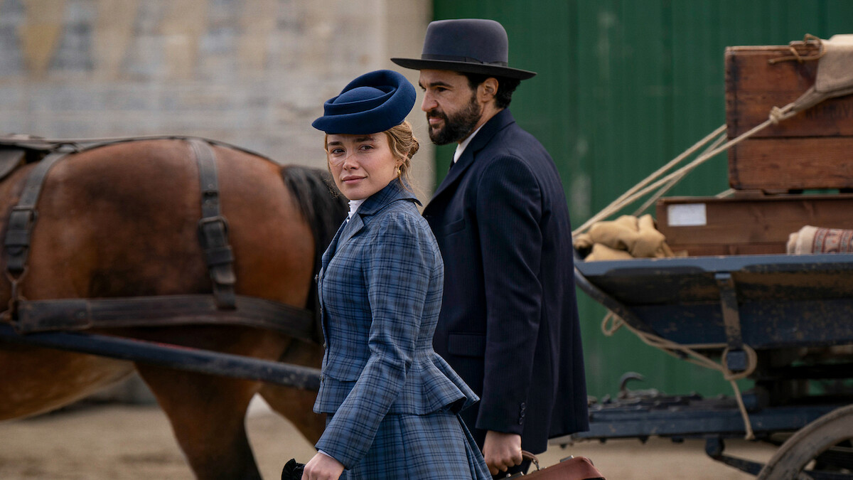 A woman and man in period clothing stand by a horse-drawn wagon with luggage, outdoors in a historical setting, suggesting a travel or arrival scene.