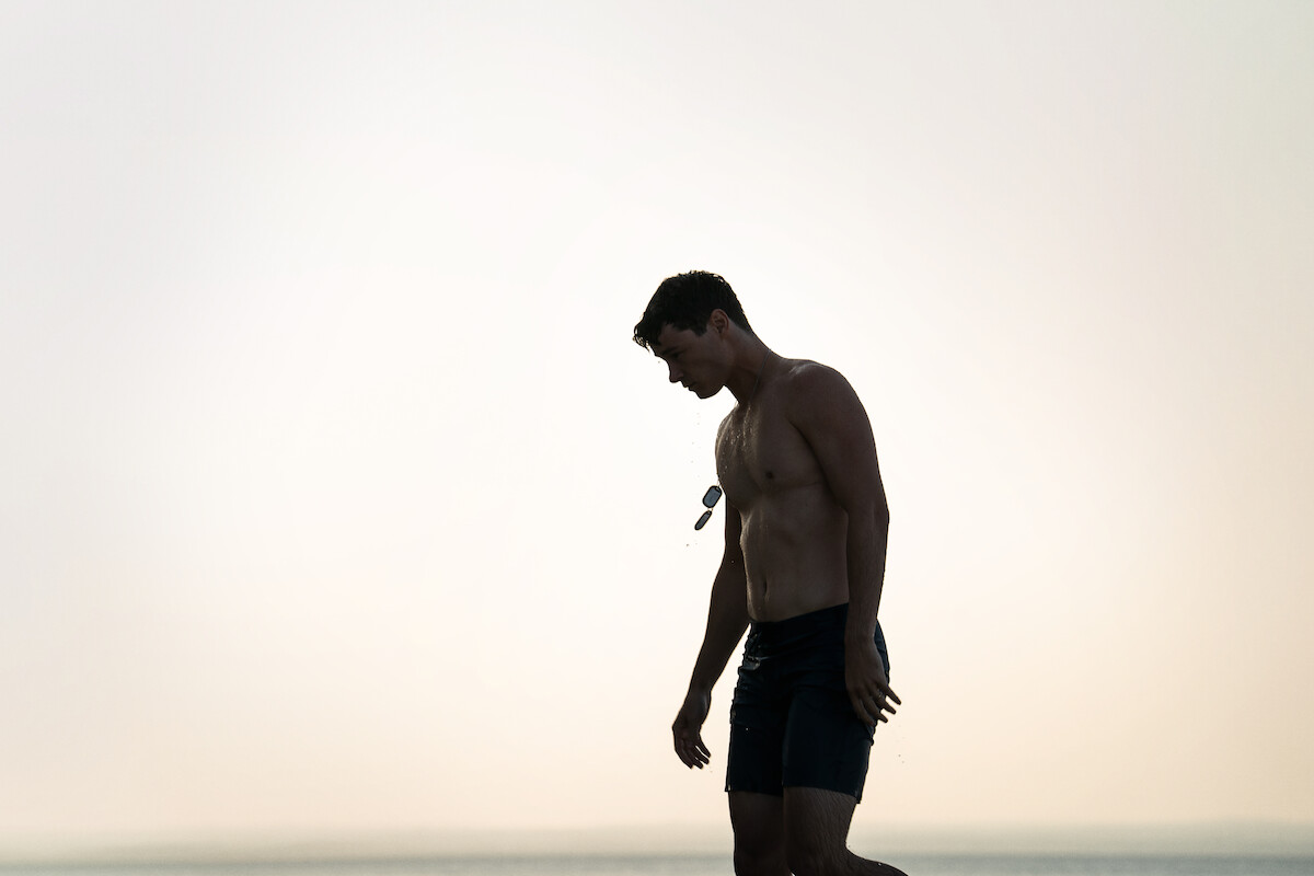 Shirtless man walking on a beach at sunset, holding sunglasses. The mood is calm and contemplative, with a soft, muted sky and ocean in the background.