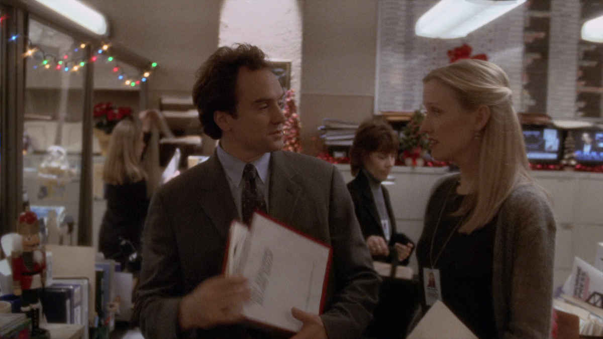 Two office workers in formal attire converse while holding documents in a decorated office with holiday lights and festive decor; other employees work in background at desks.