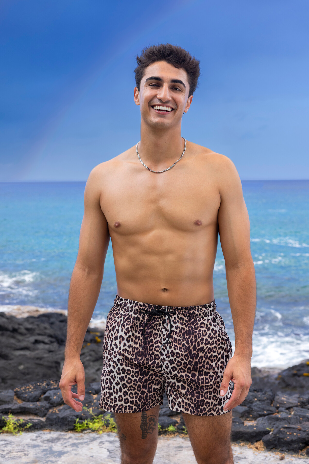 Smiling young man wearing leopard print swim trunks standing on rocky shore with blue ocean and rainbow in the background under a clear sky.