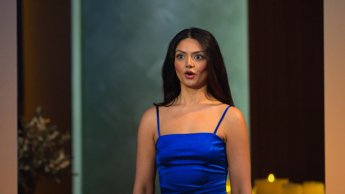 Woman in a blue dress singing or performing indoors, standing in front of a softly lit background with blurred plants and candles visible.