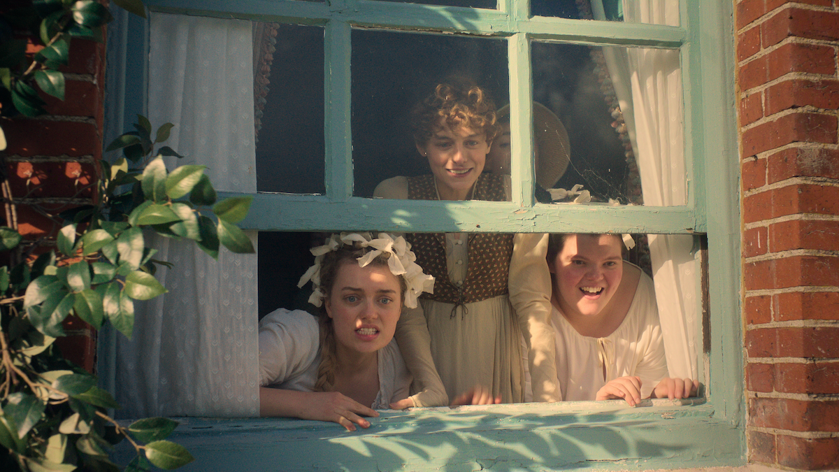 Three young women in historical clothing lean out of a blue-green window, framed by brick walls and greenery, smiling and looking outside on a sunny day. White curtains hang inside, suggesting a cozy, vintage setting.