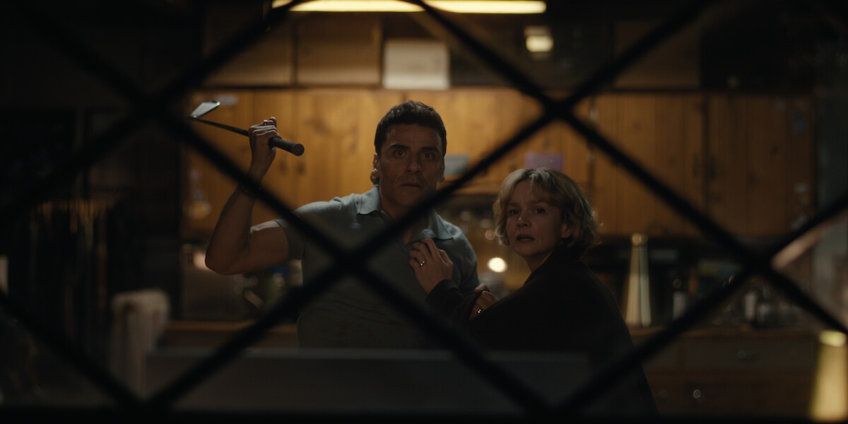 A man holding a golf club and a woman stand close together looking alarmed inside a dimly lit kitchen, viewed through a window with a crisscross frame.
