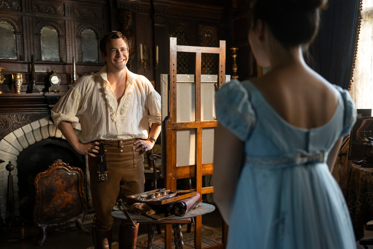 A smiling male artist stands by an easel in a richly decorated vintage room, facing a woman in a light blue dress, with painting supplies on a small table.