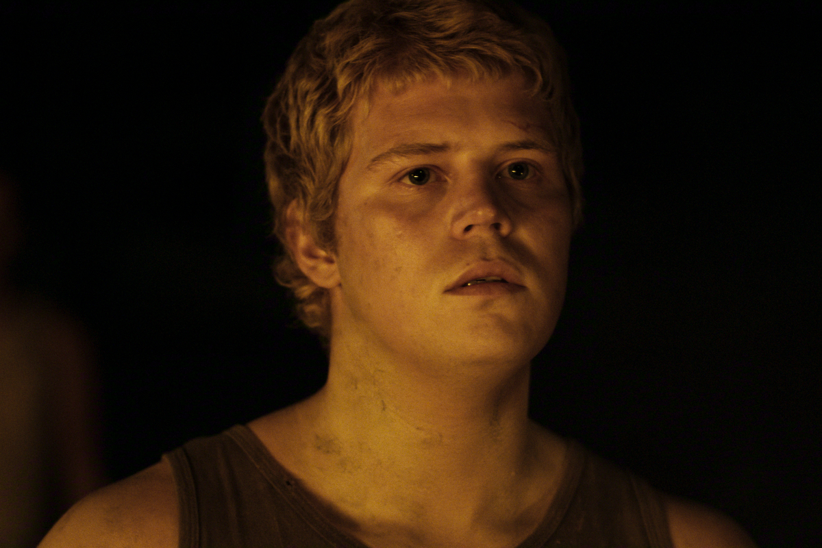 A young person with short blond hair and a dirty tank top stands in a dimly lit, dark environment, illuminated by warm, low lighting, possibly from a fire, with a concerned or thoughtful expression on their face.
