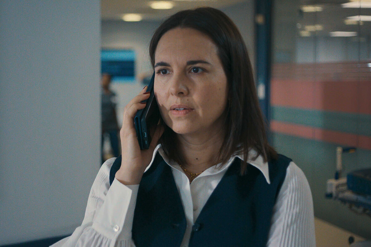 A woman with brown hair wearing a white shirt and dark vest talks on a cellphone in a modern office or hospital hallway, looking serious.