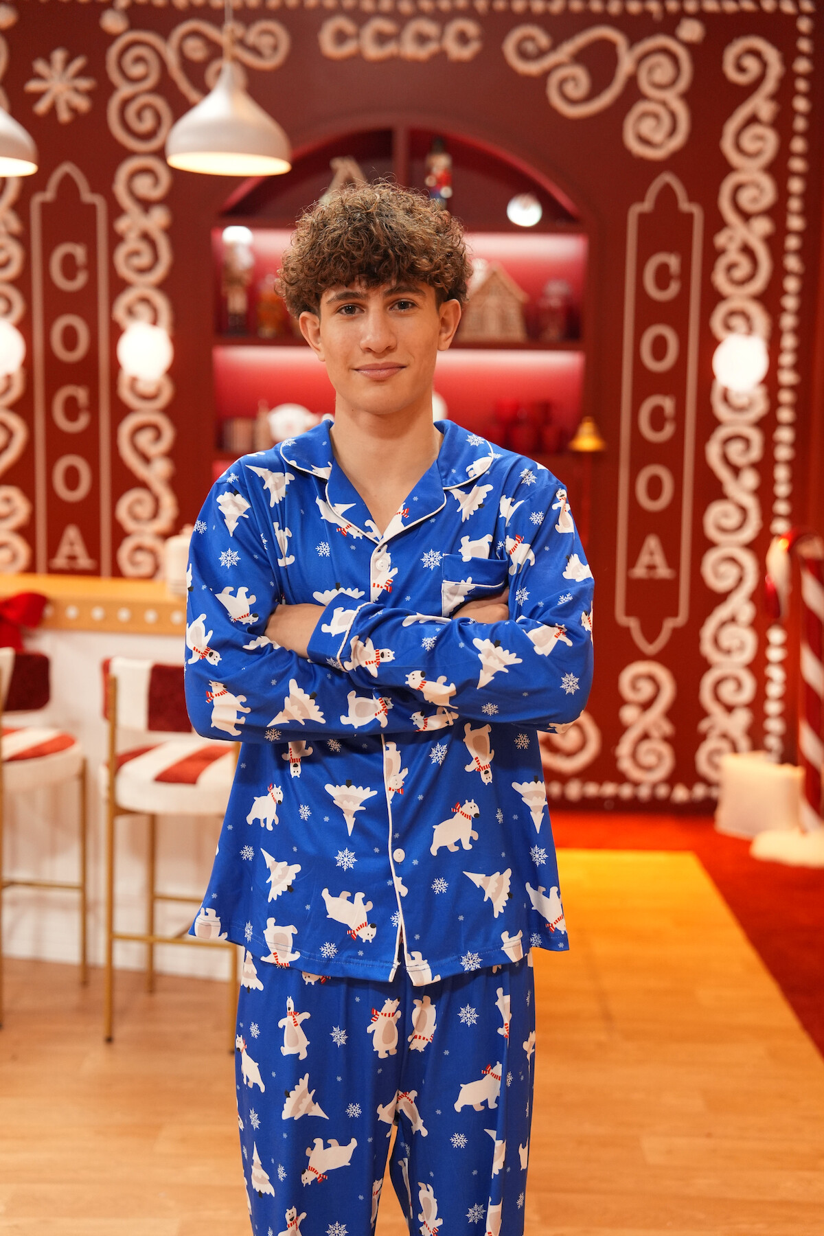 Young person in blue polar bear pajamas stands with arms crossed in a festive room decorated with red and white gingerbread patterns and holiday lights, evoking a cozy winter holiday atmosphere.