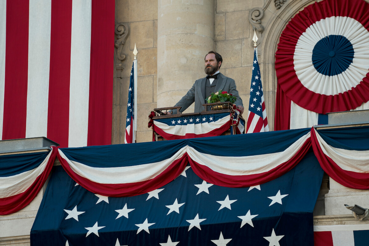 A man in historical attire stands at a decorated podium on a balcony, surrounded by American flags and patriotic bunting, evoking a formal and patriotic atmosphere.