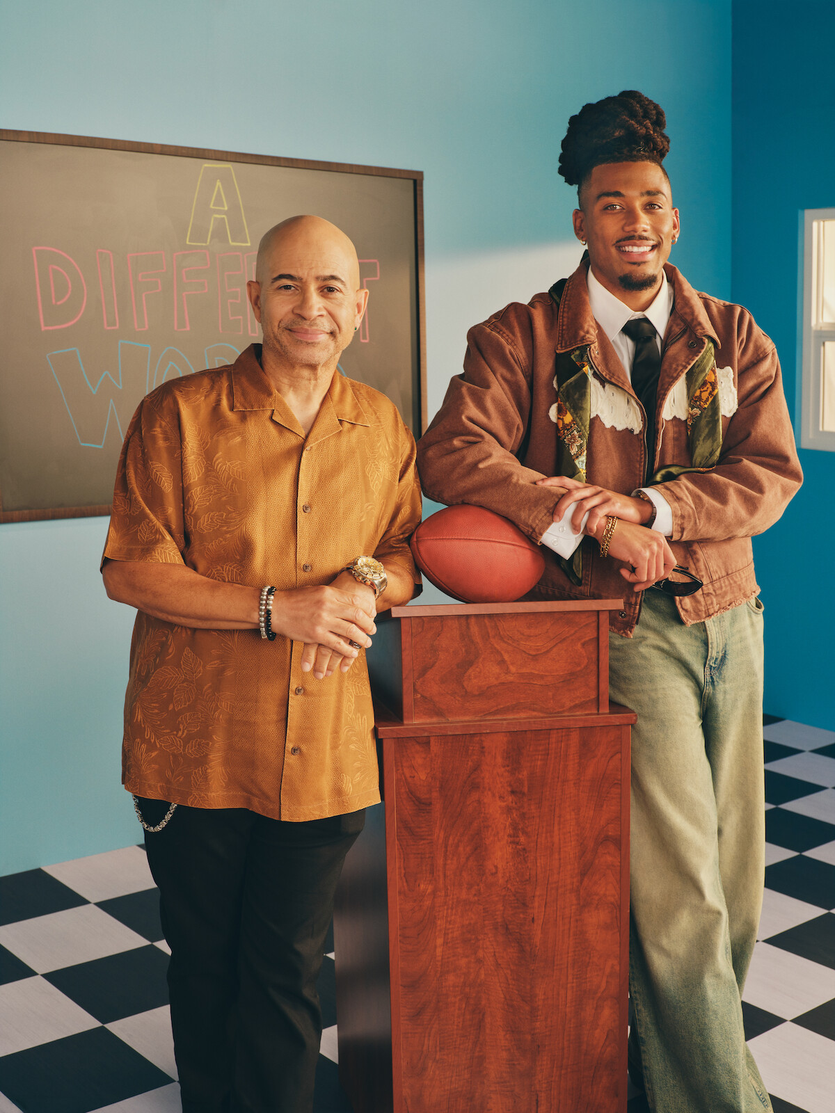 Two men stand in a brightly lit room with checkered floors; one leans on a podium holding a football, the other stands next to him. A chalkboard behind them displays the words "A Different World."